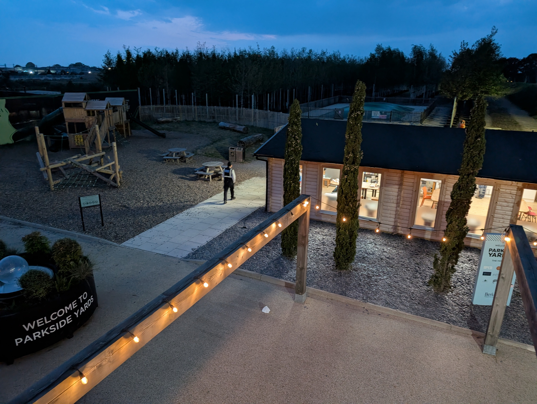 Auto-generated description: A building with a warmly lit interior is surrounded by a playground and trees in an outdoor area at dusk.